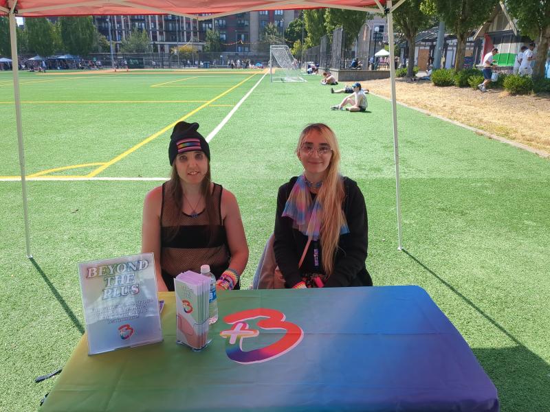 Violet Rose and Ally Kotetsu sitting at the Beyond the Plus table at the Seattle Pridefest 2025, handing out pamphlets.