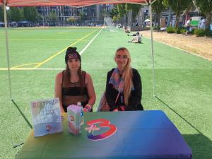 Violet Rose and Ally Kotetsu sitting at the Beyond the Plus table at the Seattle Pridefest 2025, handing out pamphlets.